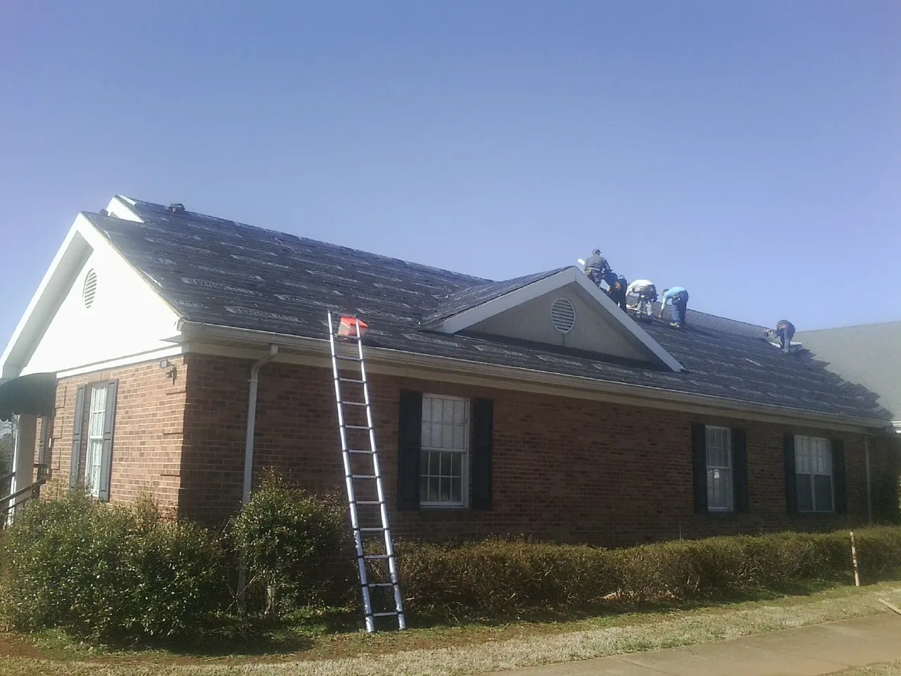 Crew tearing shingles off a brick ranch roof with a ladder set — Metal Roof Repair work by our crew in Harrells