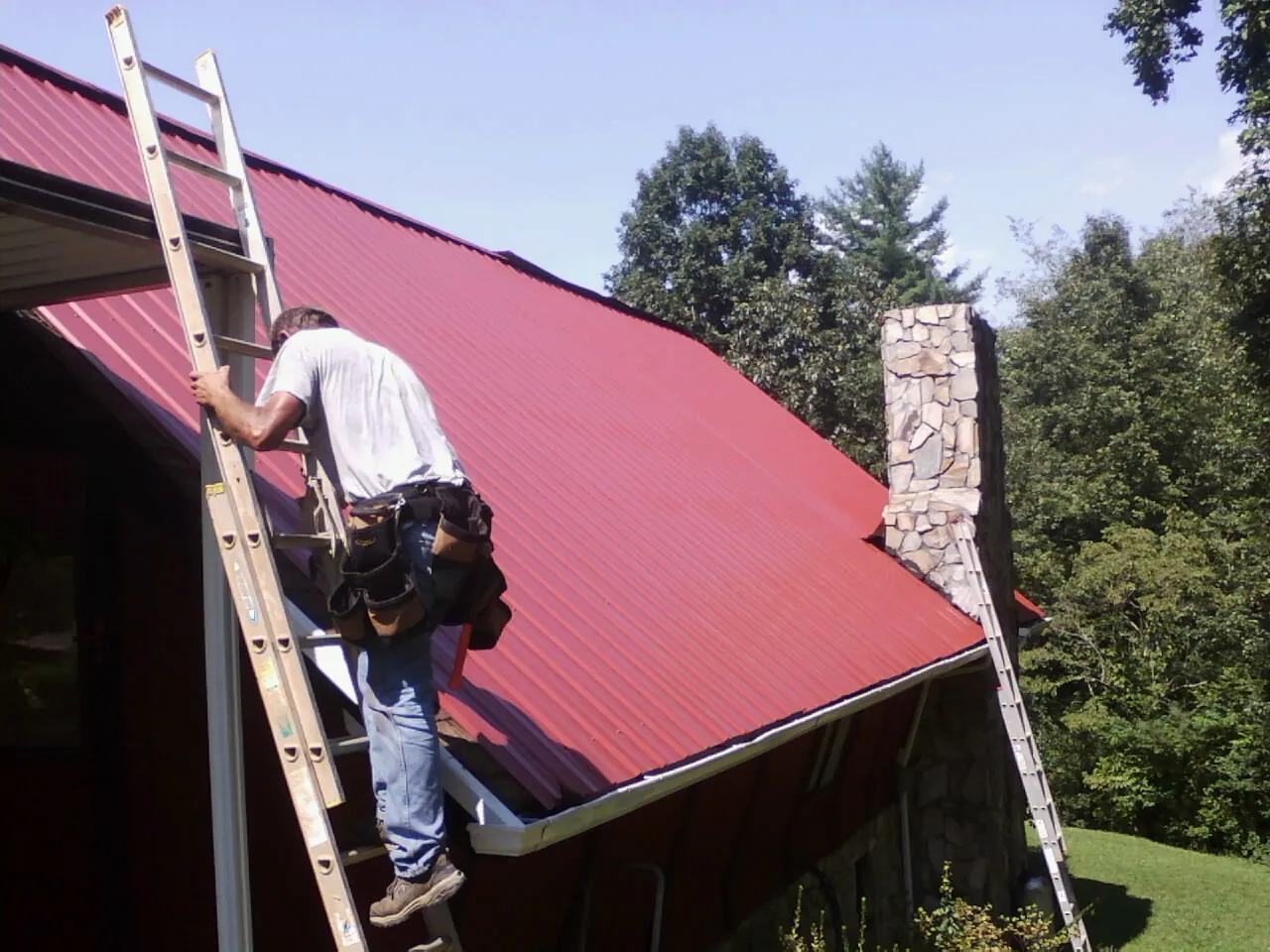 Roofer on a ladder beside a red metal roof and stone chimney — Soffit & Fascia Repair by Roofing Brothers in Harrells